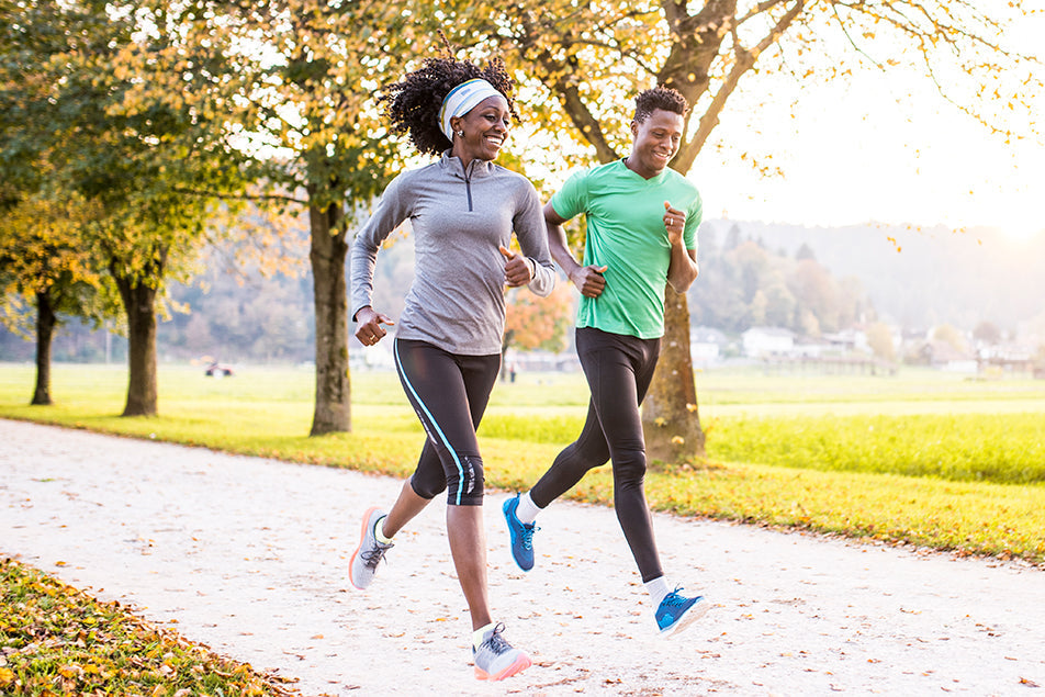 Two people jogging together on a park trail in the morning; they are smiling, wearing athletic clothing, and surrounded by green grass and trees with autumn leaves