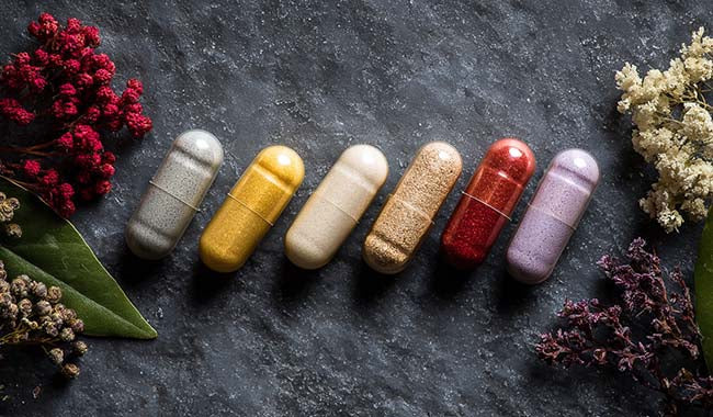 Six colorful supplement capsules arranged on a dark surface with flowers around the edges